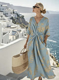 Woman wearing a flowing blue botanical print tie-waist maxi dress while overlooking the ocean at a coastal resort. Woman in a light blue dress with gold patterns standing on a balcony overlooking a scenic coastal town.