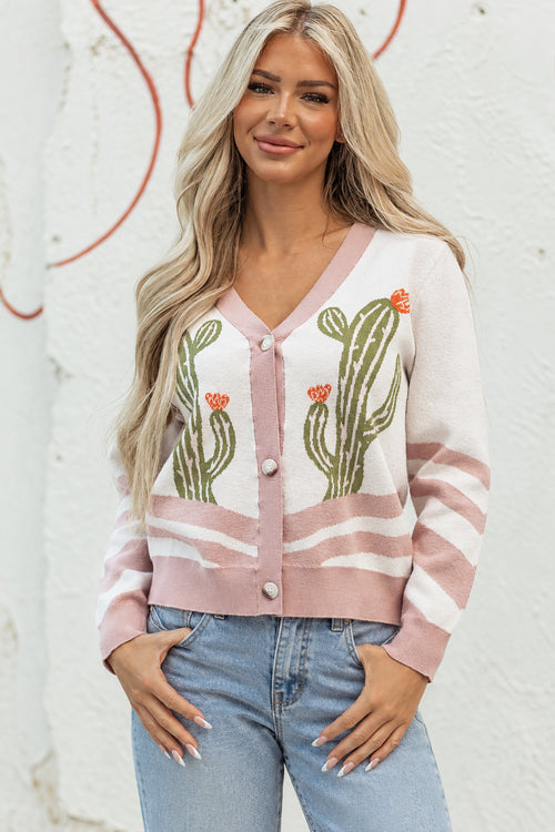 A woman modeling a pink and white knit cardigan with green cactus and orange flower motifs, paired with dark denim shorts against a textured white wall.
