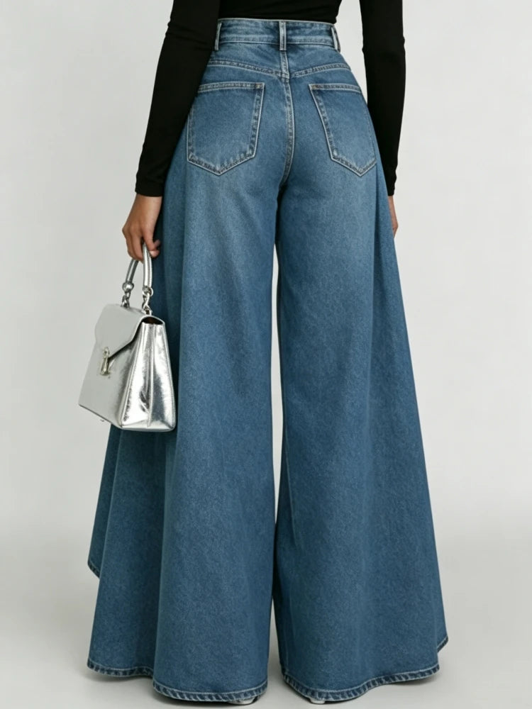 A person shown from the back wearing high-waisted, wide-leg blue denim jeans with a black long-sleeve top and holding a metallic silver handbag against a light gray studio background.