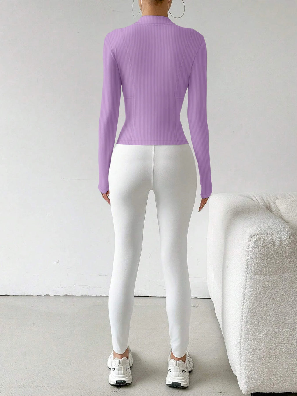 A woman shown from the back wearing a fitted long-sleeve ribbed top in light purple and white leggings, standing in a minimalist room with a white textured sofa on the right.