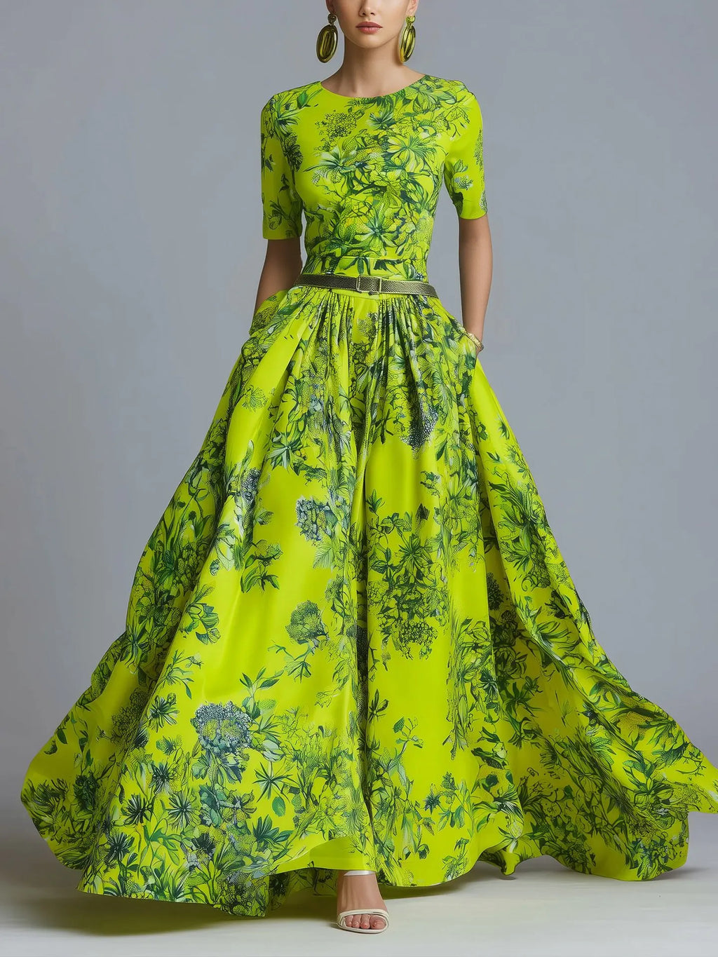 A model wearing a vibrant lime green and dark green floral print maxi dress with a fitted bodice, short sleeves, and a full flowing skirt, styled with large gold hoop earrings and clear high-heeled sandals against a neutral gray background.