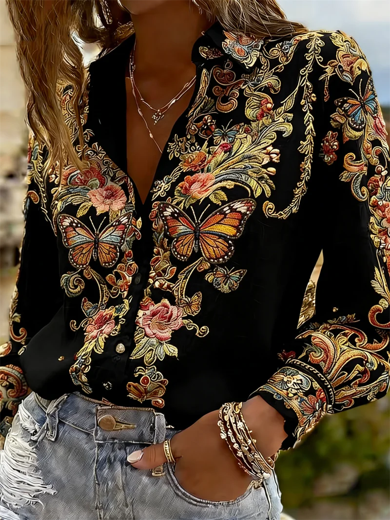 A close-up of a woman wearing a black long-sleeve button-up blouse with ornate multicolor butterfly and floral embroidery, paired with light-wash distressed denim shorts.