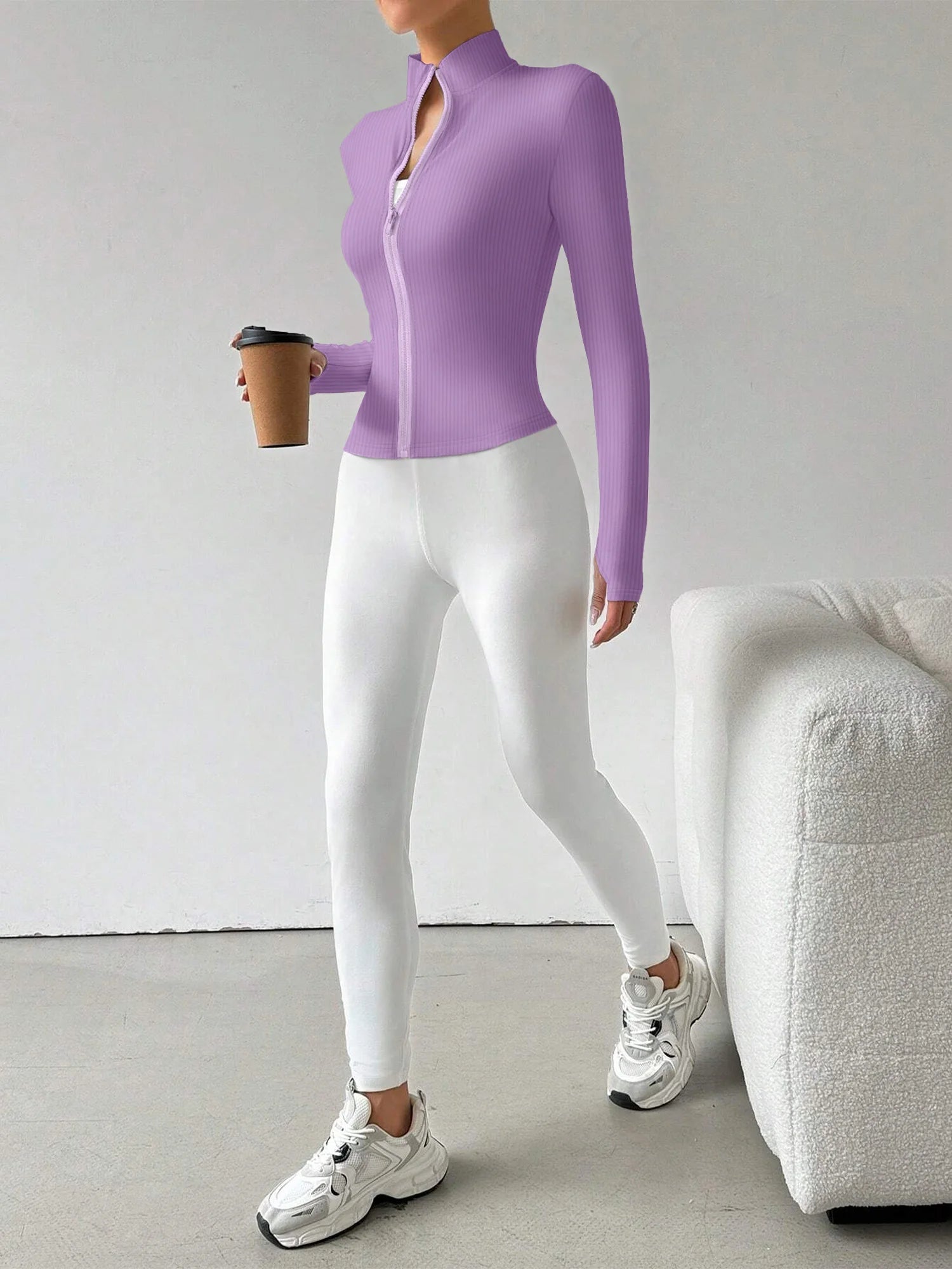 A woman modeling a fitted long-sleeve zip-front athletic jacket in light purple, paired with white leggings and white sneakers, standing indoors and holding a takeaway coffee cup.