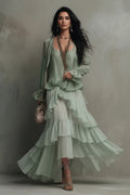 A woman models a light sage green two-piece outfit consisting of a long-sleeve blouse with ruffled details and a matching tiered ruffle maxi skirt, styled with a long statement necklace and holding a small metallic clutch against a textured gray wall.
