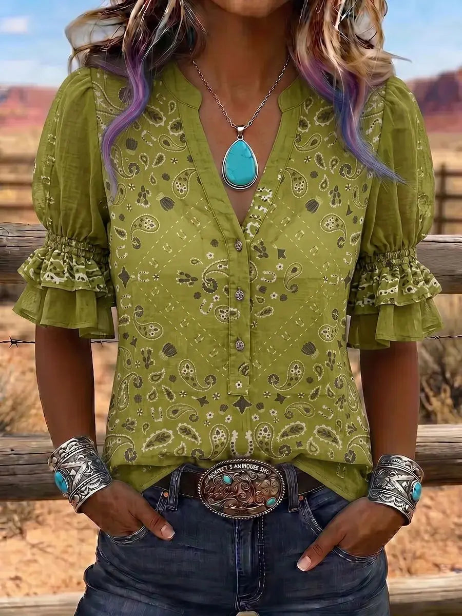 A woman wearing a sage green bohemian-style blouse with a paisley pattern, puff sleeves, and a V-neckline, paired with a turquoise pendant necklace and silver bracelets, set against a rustic outdoor background.