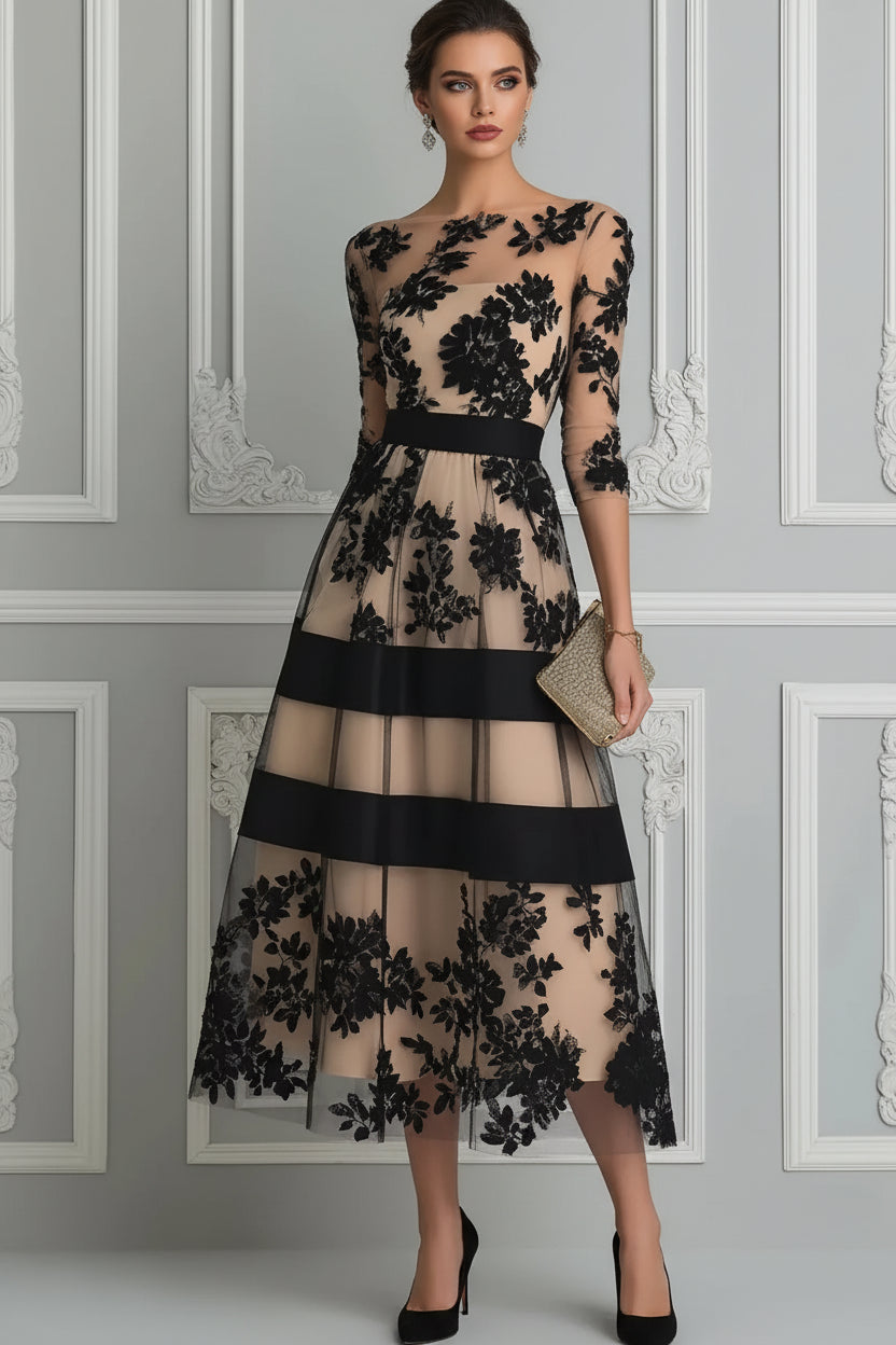 A woman models a sheer beige midi dress with black floral lace applique and wide black horizontal bands, holding a small metallic clutch against a decorative paneled wall.