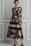 A woman models a sheer beige midi dress with black floral lace applique and wide black horizontal bands, holding a small metallic clutch against a decorative paneled wall.