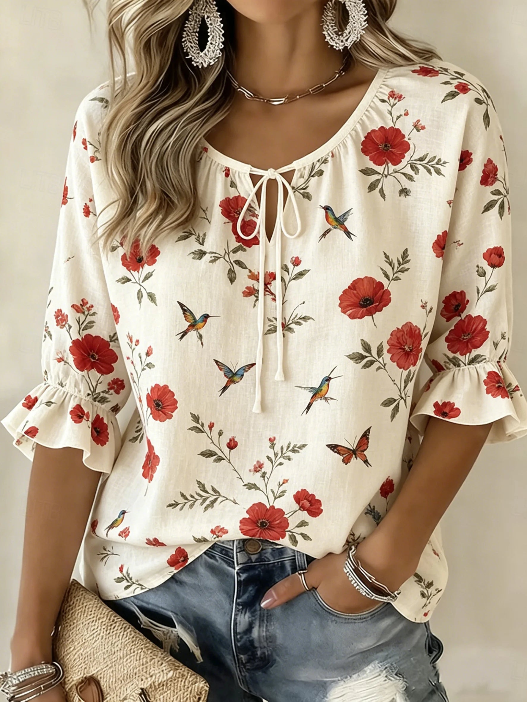 A woman wearing a cream-colored short-sleeve blouse with a red floral and bird print, paired with distressed blue jeans and silver jewelry.
