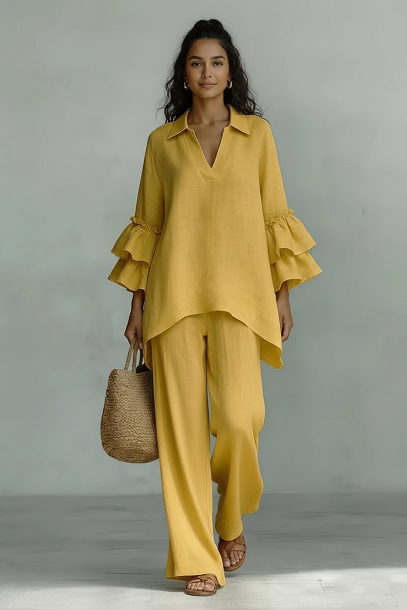 A woman wearing a loose, two-piece yellow linen outfit with a ruffled long-sleeve top and wide-leg pants, holding a woven straw bag against a neutral gray studio background.