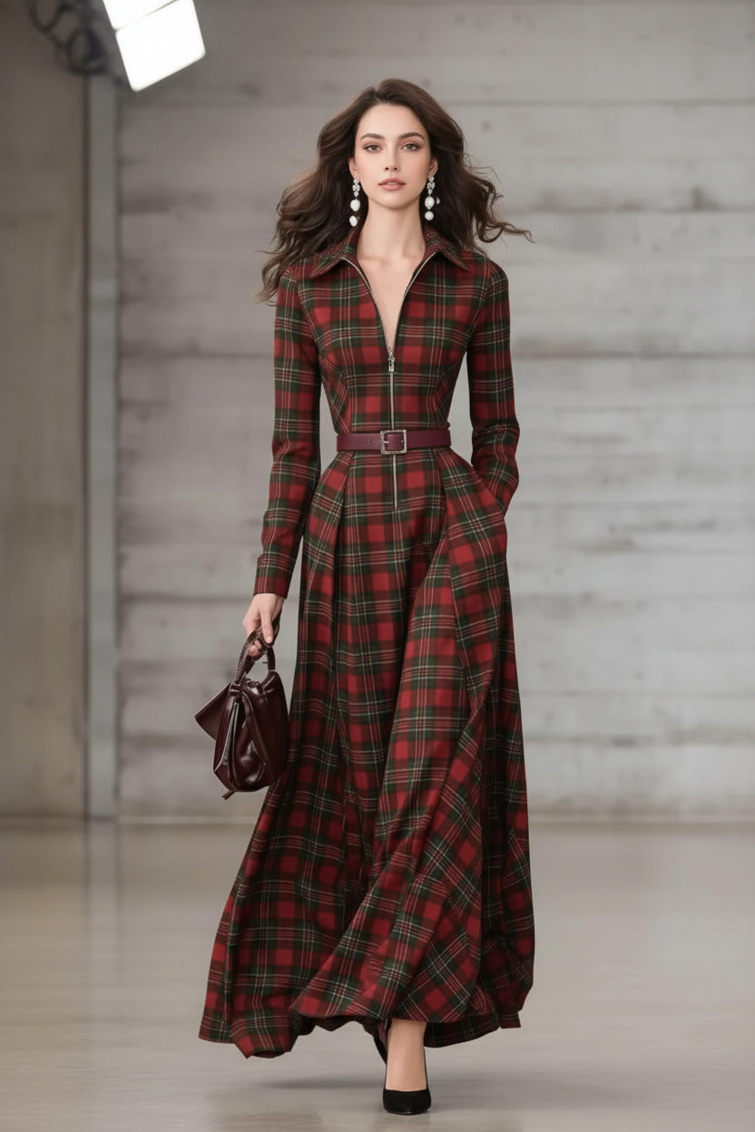 A woman models a long-sleeve plaid maxi dress with a collar, front zipper, and matching belt. She is holding a dark brown handbag against a neutral gray concrete wall background.