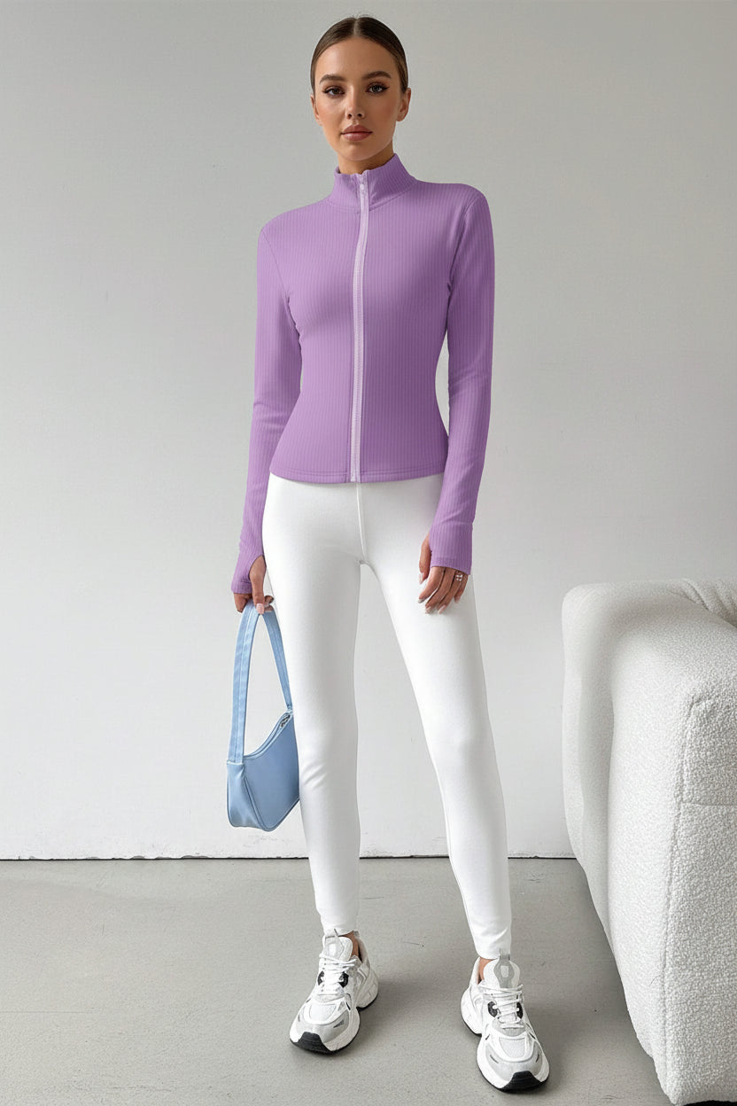 A model wearing a fitted long-sleeve zip-up athletic top in light purple, paired with white leggings and white sneakers, standing in a minimalist room next to a white textured sofa.