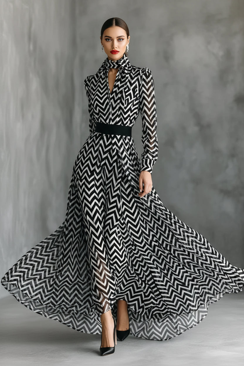A woman models a long-sleeve high-neck maxi dress with a bold black and white chevron pattern and a black waist belt, posed against a textured gray studio backdrop.