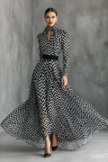 A woman models a long-sleeve high-neck maxi dress with a bold black and white chevron pattern and a black waist belt, posed against a textured gray studio backdrop.