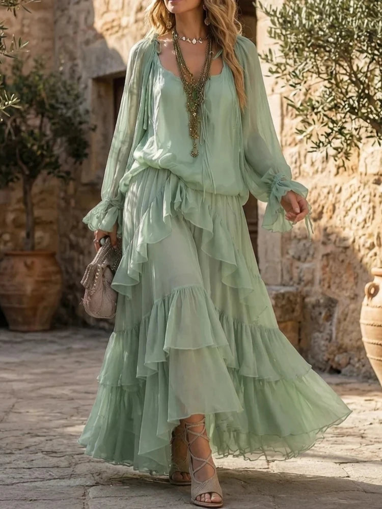 A woman wearing a flowing sage green bohemian maxi dress with a sheer long-sleeve layer, ruffled layers, and a tiered skirt, standing in a rustic stone courtyard.