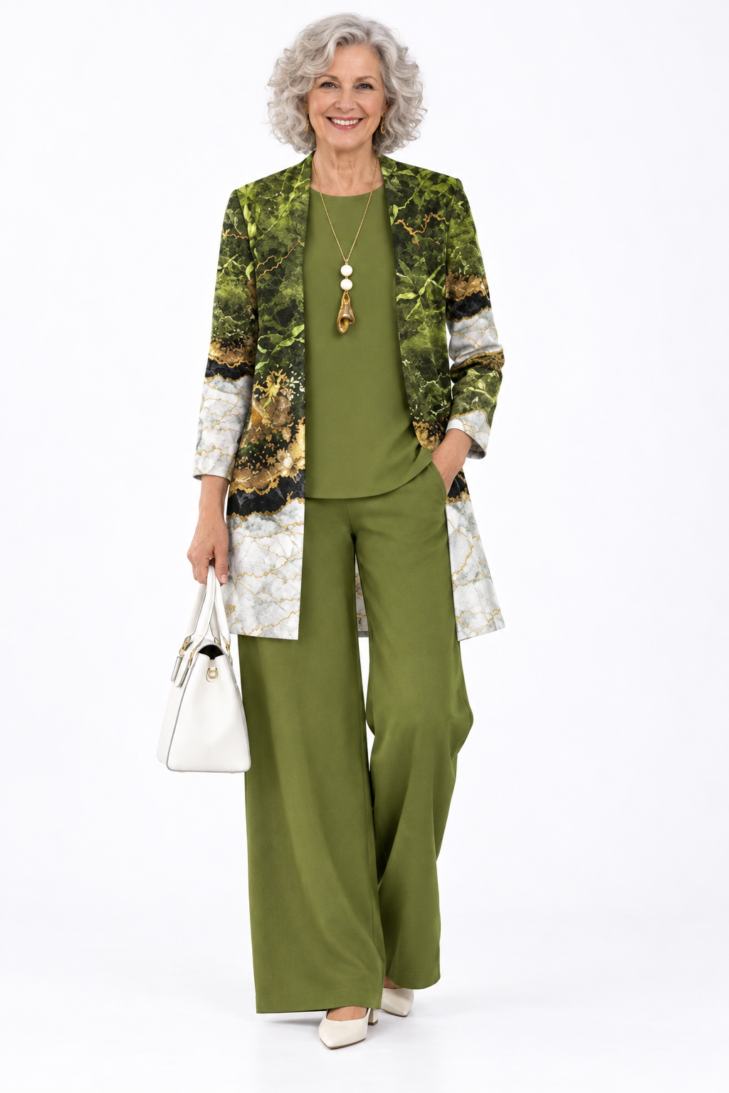 An older woman stands against a plain white studio background wearing a coordinated outfit consisting of an olive green top and wide-leg pants with a green-and-white marble-patterned blazer. She is holding a white handbag and wearing a long pendant necklace.