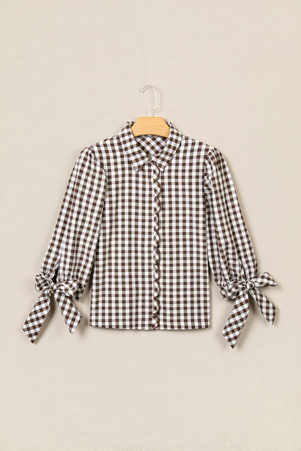 A long-sleeve button-front blouse with a brown and white gingham check pattern, featuring tie details on the sleeves, displayed on a wooden hanger against a light beige background.